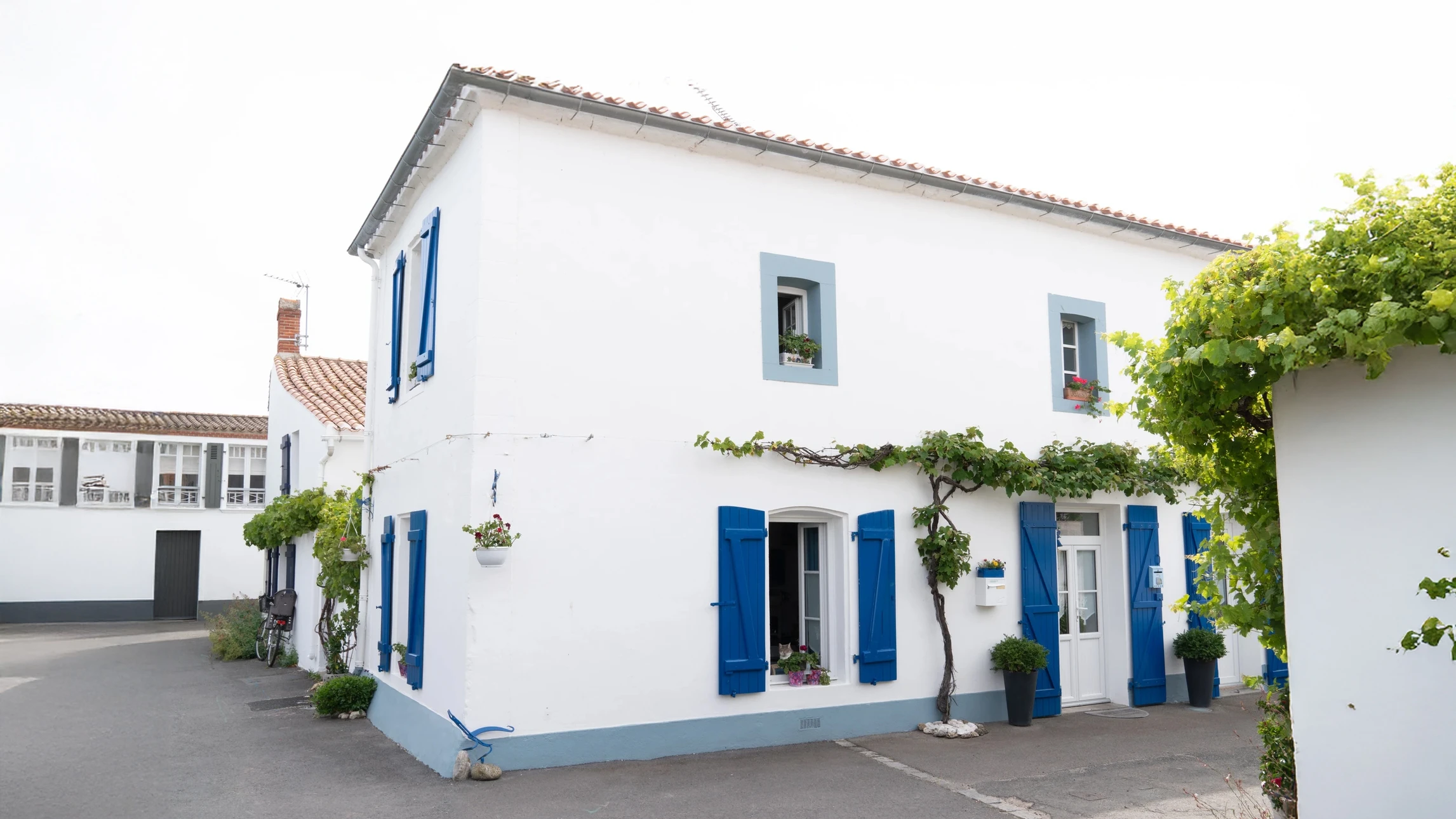 Façade de maison traditionnelle française aux murs blancs avec volets bleus ouverts, porte-fenêtre centrale et fenêtres à l'étage